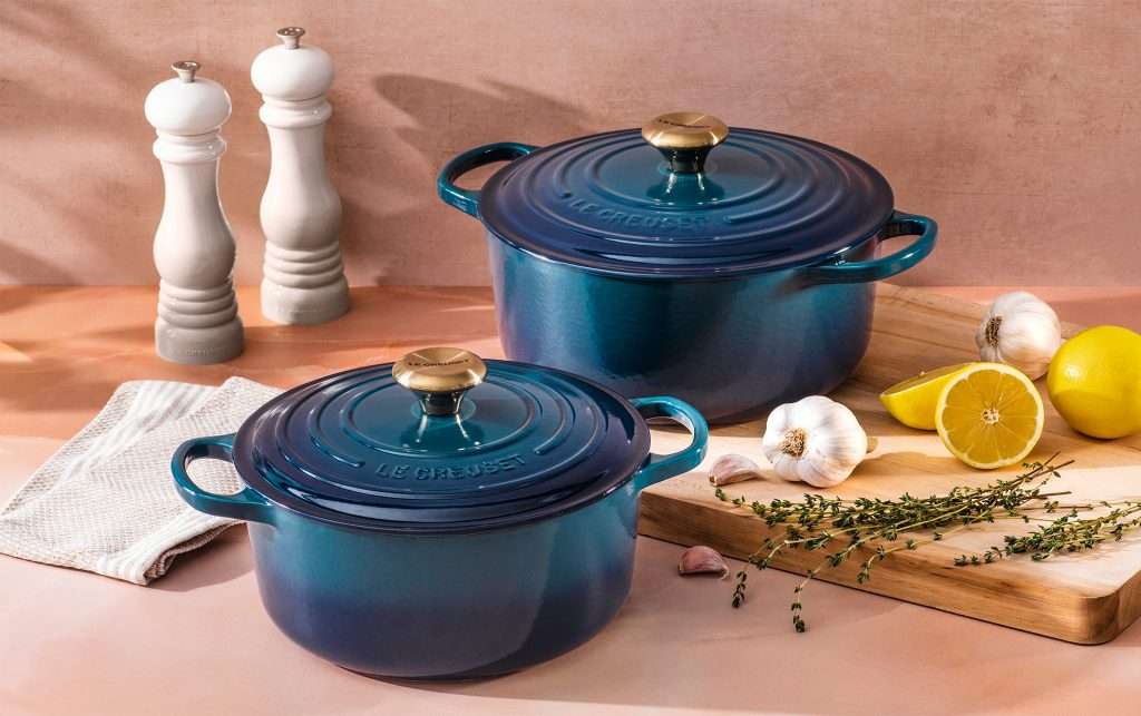 Two blue Le Creuset pans on table with pepper and salt and cutting board