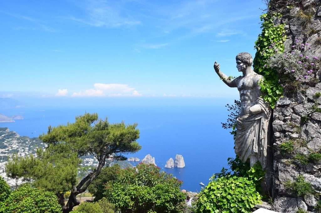 The Island of Capri with a view over the sea with a roman statue. In the background the dramatic Faraglioni, three towering rock formations which jut out from the Mediterranean just off the island's coast.